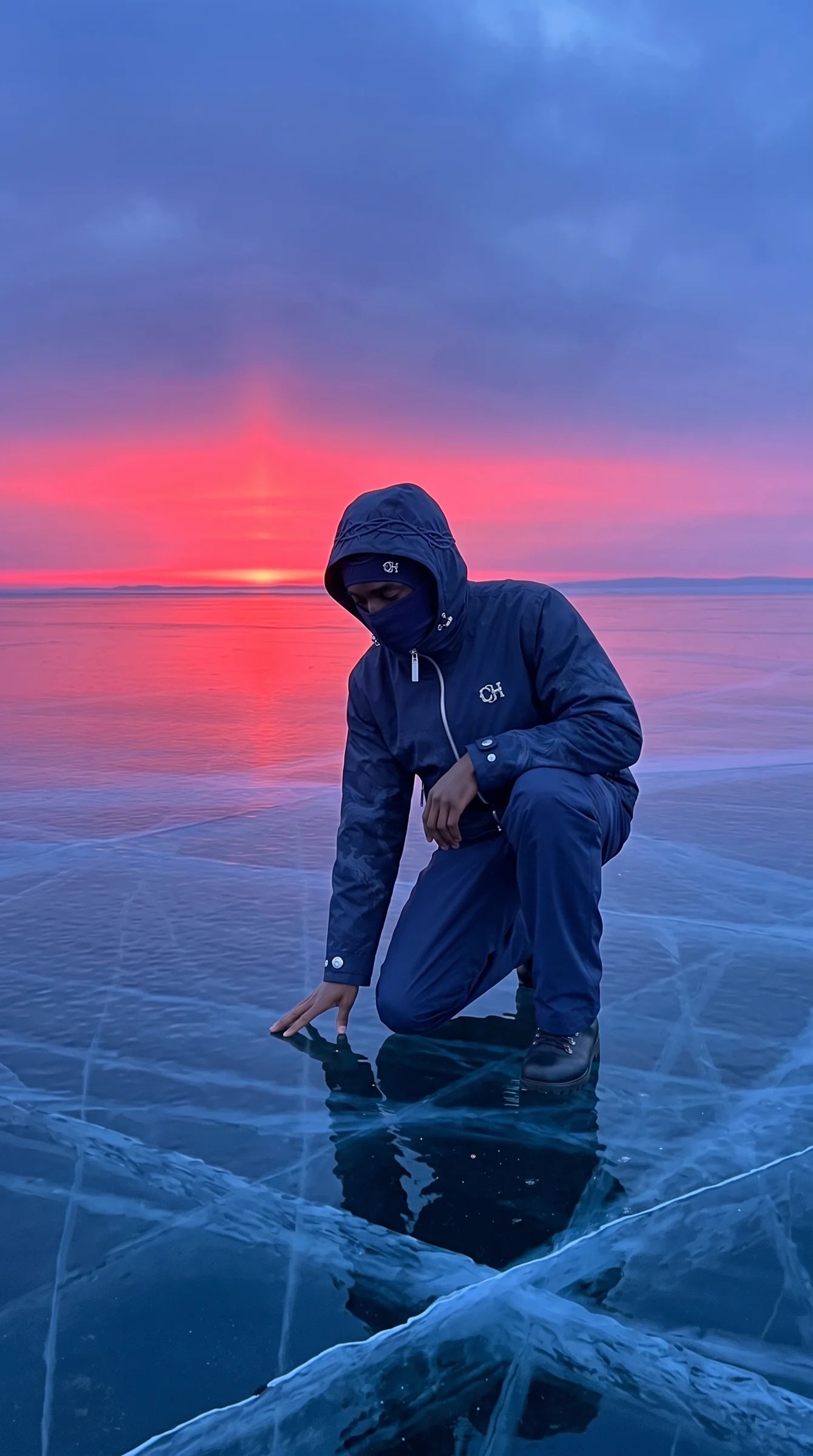The navy EXODUS Rain Shell, kneeling on a frozen sea at sunrise.